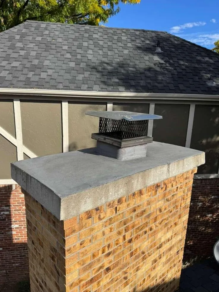 A light orange brick chimney with a gray concrete crown and a black mesh spark-screen cap beside a house with a dark shingle roof