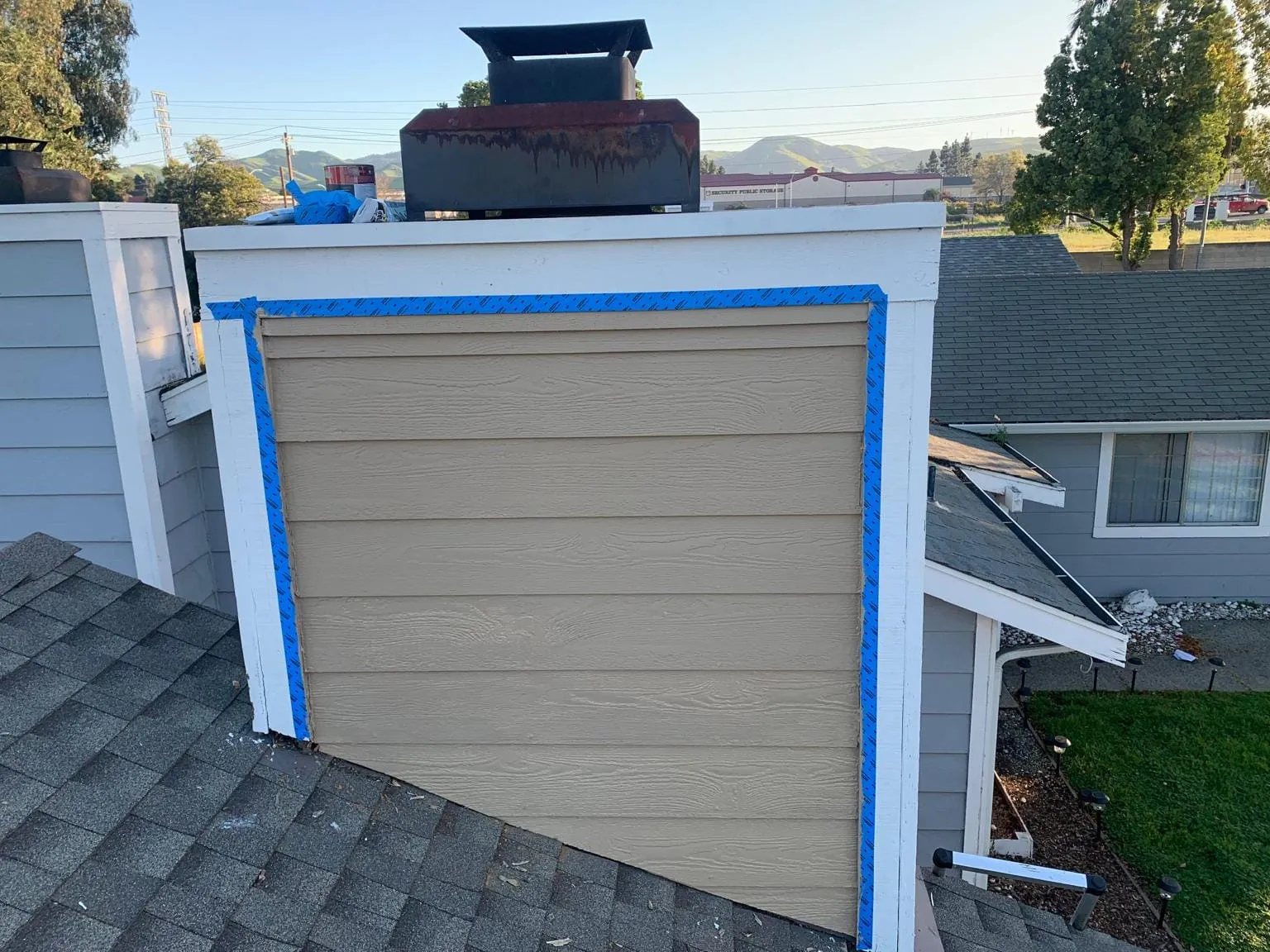 A black cap on top of a tan siding-covered chimney chase trimmed in white and blue tape, with low hills and a sunny sky behind