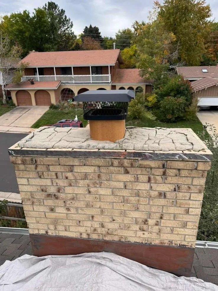 A flat-topped tan brick chimney with a cracked concrete crown and a small black cap, above a street of two-story houses and autumn trees