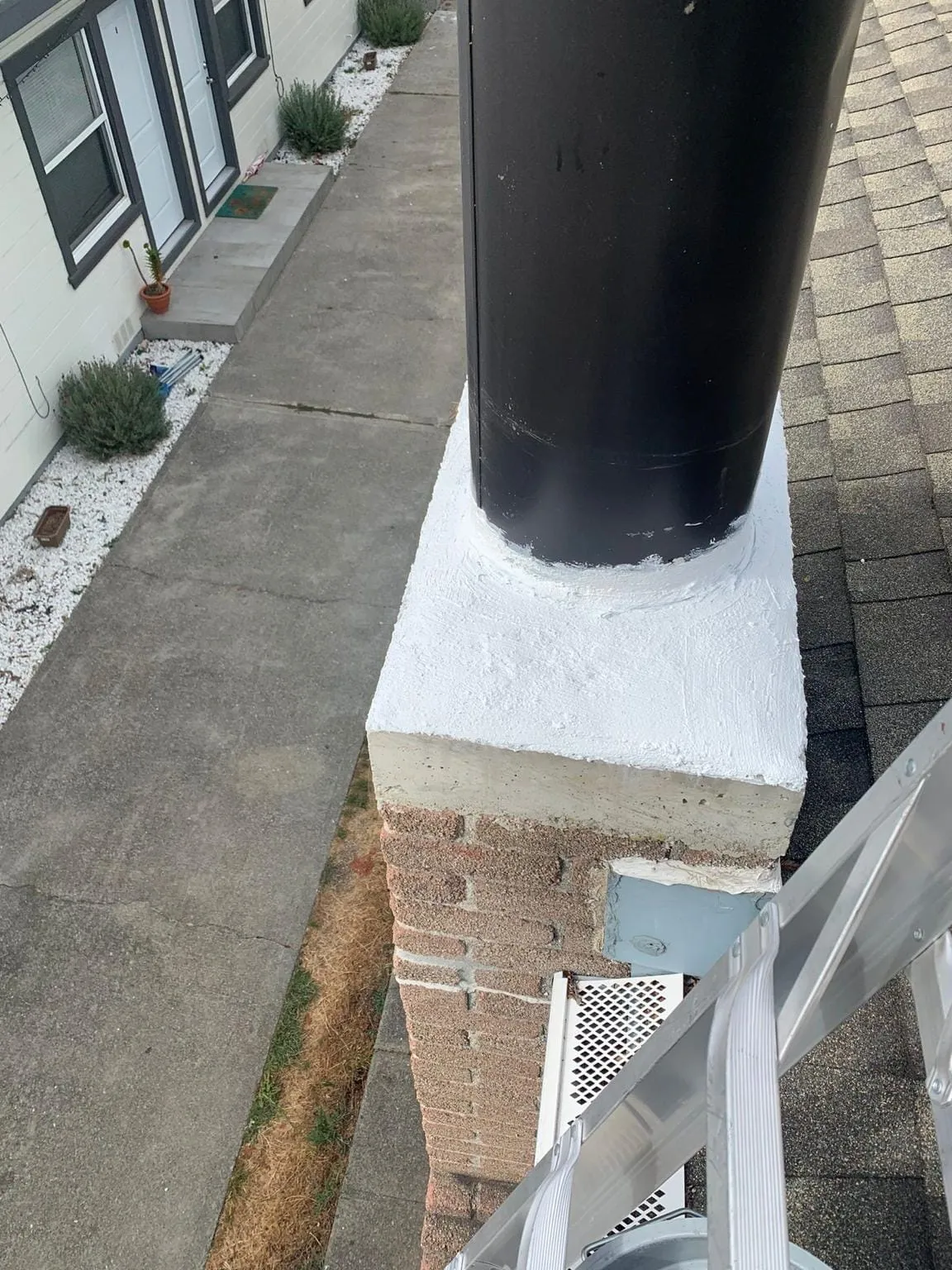 A black flue pipe rising from a brick chimney whose top is coated in white sealant, beside an aluminum ladder and a concrete path between two buildings