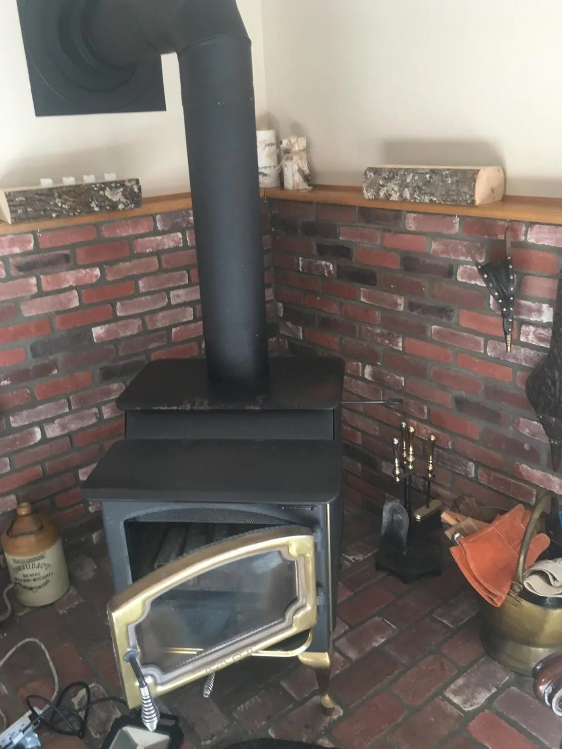 A black wood stove with a glass door ajar and a tall stovepipe against a corner of dark red brick, with fireplace tools beside it